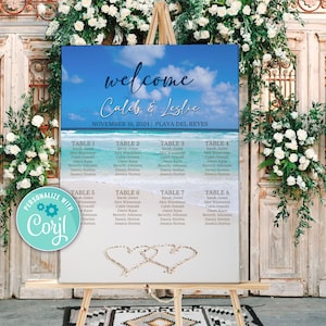 May include: A wedding seating chart with a beach theme. The chart is on a wooden easel and features a blue and green ocean scene with white clouds. The text "welcome Caleb & Leslie" is at the top of the chart. The chart lists the names of guests and their assigned tables.