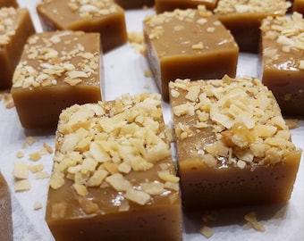 May include: Close-up of several square caramel candies topped with chopped nuts. The candies are a light brown color and sit on a white surface. The nuts are a lighter shade of brown and add texture to the smooth caramel.