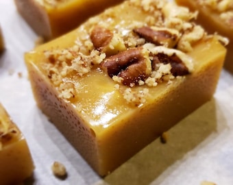 May include: Close-up of a square piece of caramel fudge topped with chopped pecans. The fudge is a light golden brown color and has a glossy finish.