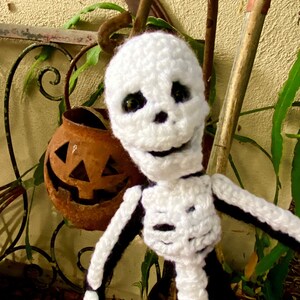 May include: White crocheted skeleton doll with black details, smiling with large black eyes. The doll is posed with one arm raised and the other arm extended. The doll is in front of a rusty pumpkin and a brown wooden stick.