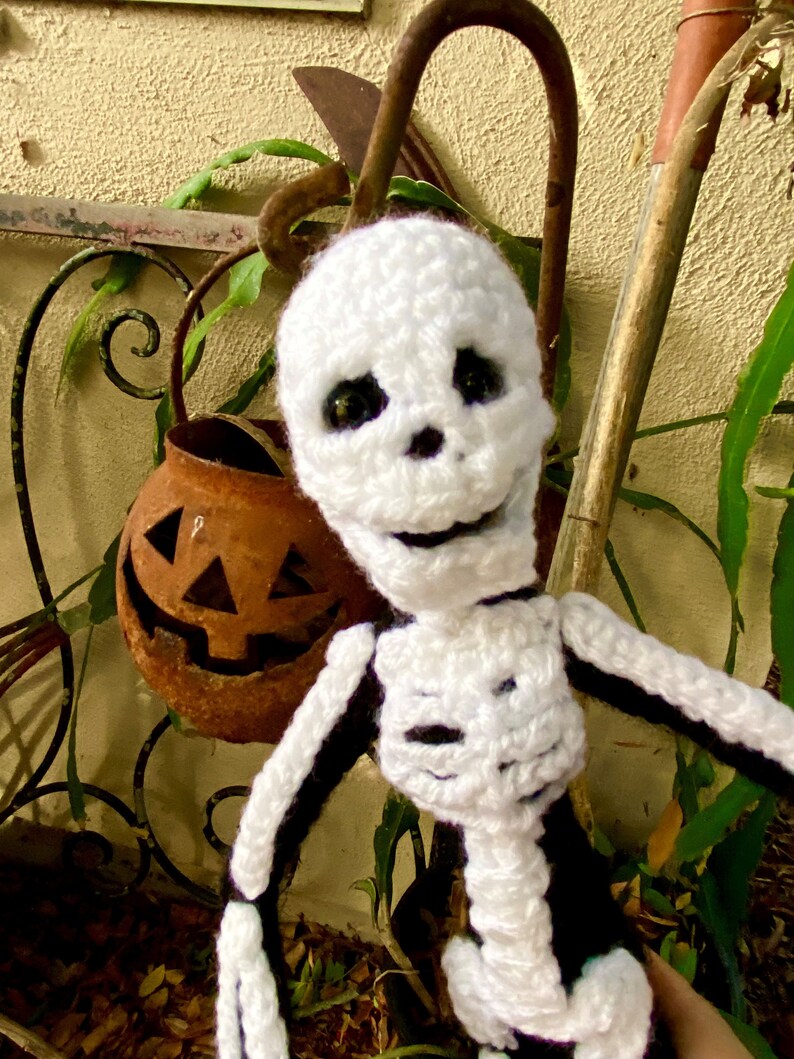 May include: White crocheted skeleton doll with black details, smiling with large black eyes. The doll is posed with one arm raised and the other arm extended. The doll is in front of a rusty pumpkin and a brown wooden stick.