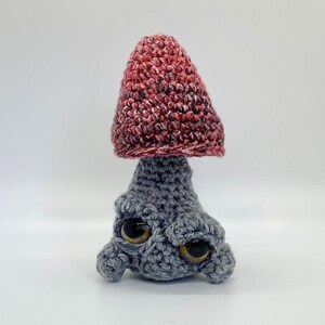 May include: A crocheted mushroom toy with a red and white cap and a gray body. The mushroom has large, black eyes and a whimsical expression.