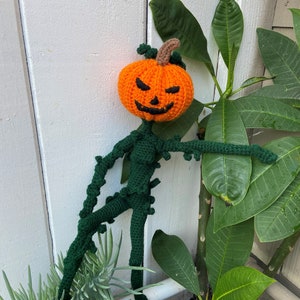 Puede incluir: Una calabaza de crochet con un cuerpo de vid verde y una cara de miedo. La calabaza es naranja con ojos negros y una boca. El cuerpo de la vid es verde y tiene un tallo largo y delgado. La calabaza está apoyada en una pared blanca y una planta verde.