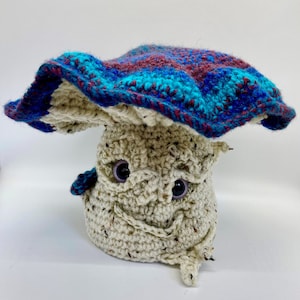 May include: A crocheted mushroom toy with a blue, purple, and red patterned cap. The mushroom has a white body with a smiling face and two black eyes.