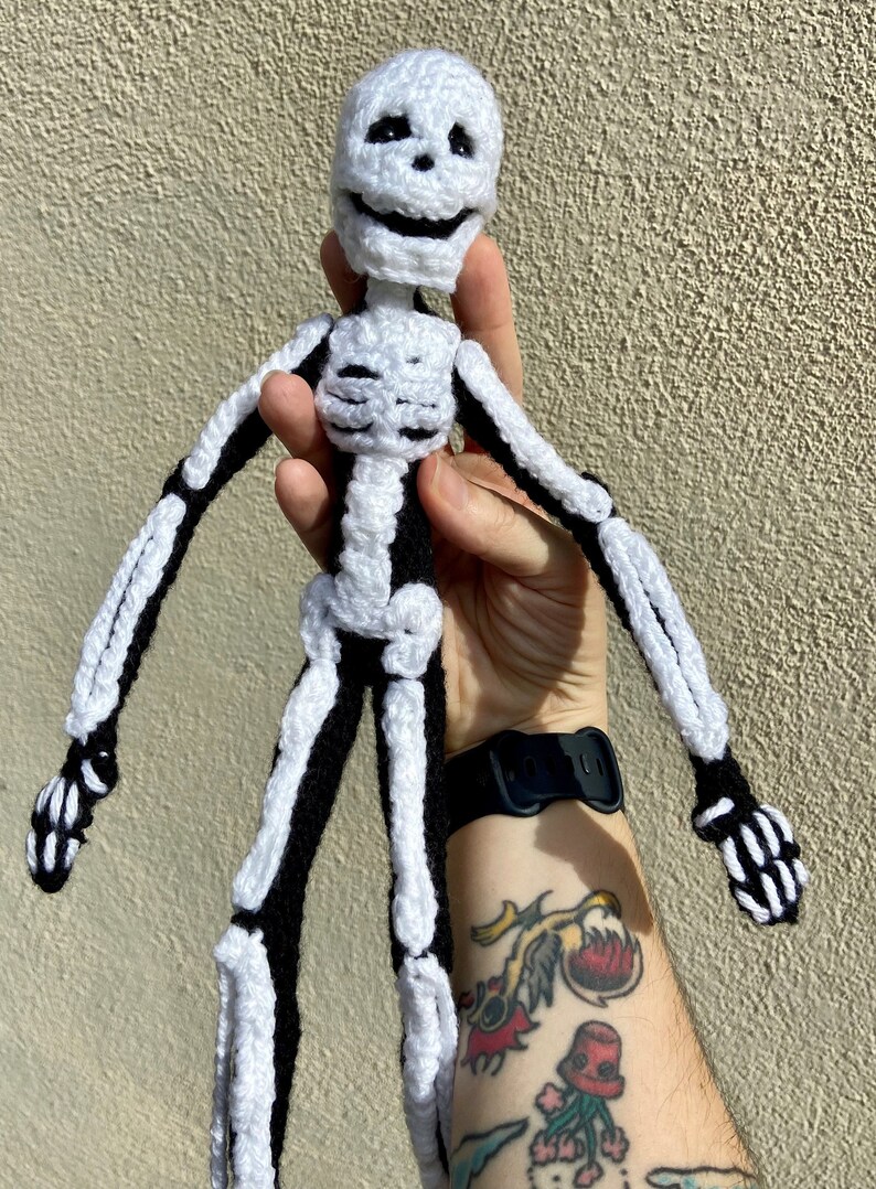 May include: A crocheted skeleton doll with black and white yarn. The skeleton is smiling and has a friendly expression.