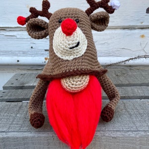 May include: A crocheted reindeer gnome with a red beard, brown body, and white face. The gnome has a red nose and antlers with a white pom-pom on one antler.