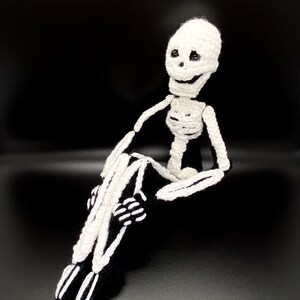 May include: A crocheted skeleton doll with a white body and black bones. The skeleton is sitting with its legs crossed and has a friendly expression.