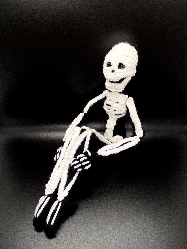 May include: A crocheted skeleton doll with a white body and black bones. The skeleton is sitting with its legs crossed and has a friendly expression.
