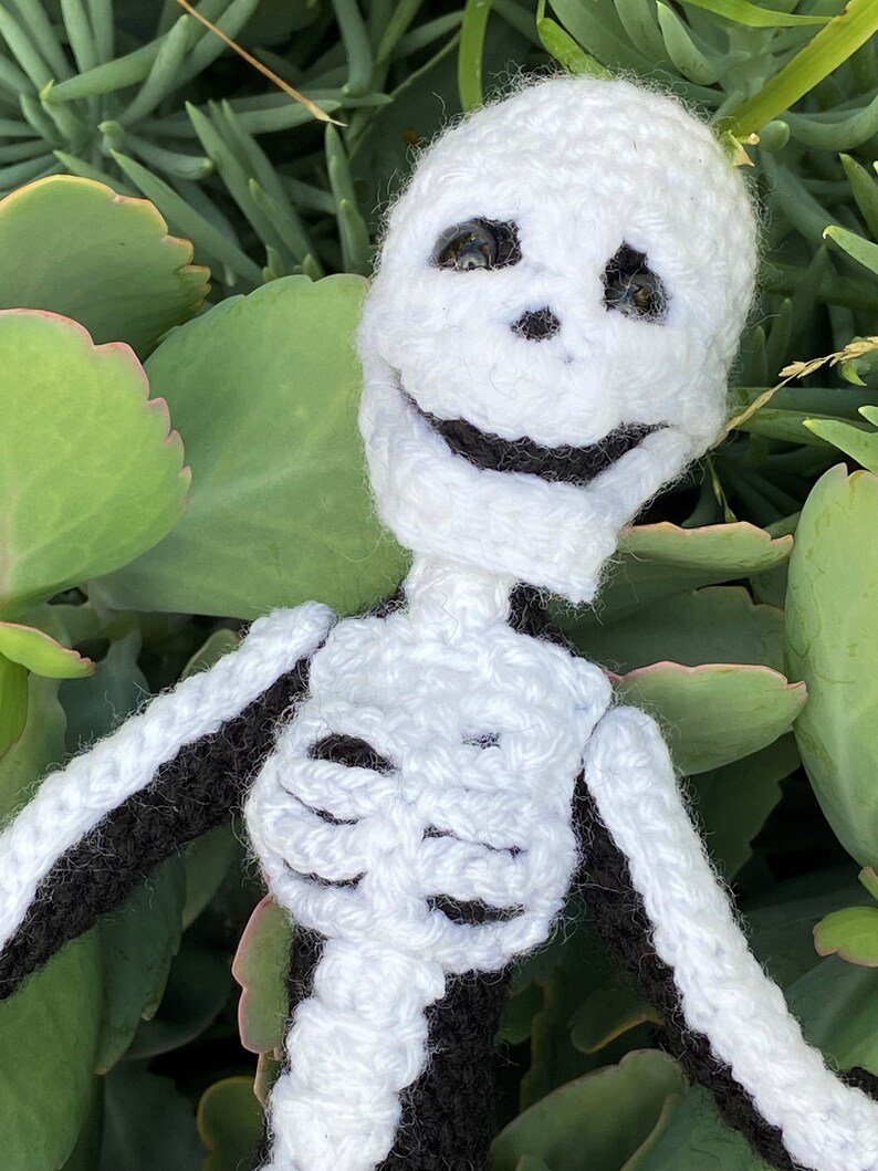 May include: A crocheted skeleton doll with a white body and black arms and legs. The skeleton has a smiling face with black eyes and a black mouth.
