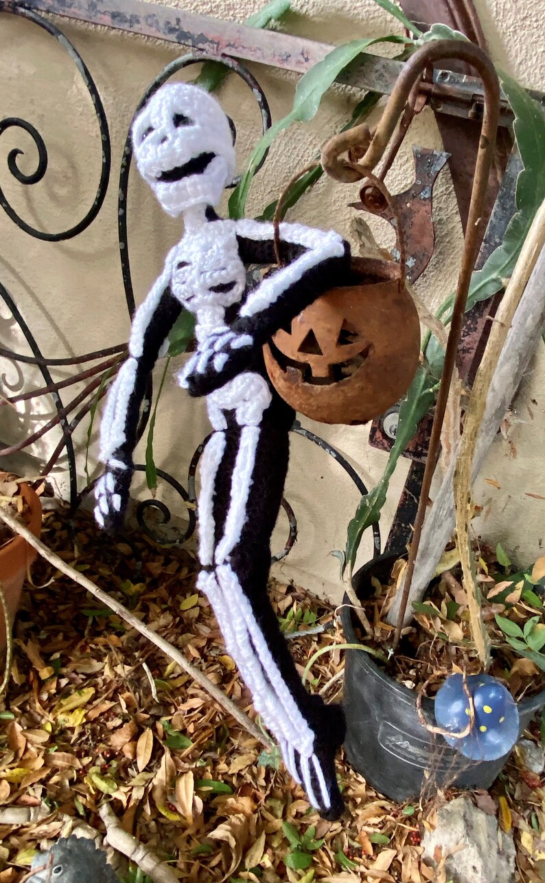 May include: A crocheted skeleton doll holding a carved pumpkin. The doll is wearing a black and white skeleton costume. The pumpkin is brown and has a carved jack-o'-lantern face.