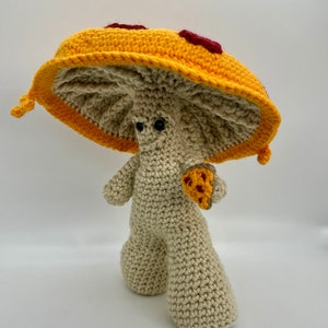 May include: A crocheted mushroom toy with a pizza-shaped cap. The cap is yellow with red pepperoni and a smiling face. The mushroom is holding a small pizza slice in its hand.