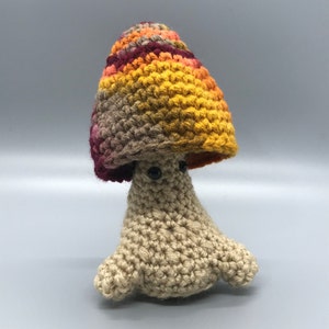 May include: A crocheted mushroom toy with a brown, red, and yellow striped hat. The mushroom has a beige body and black eyes.