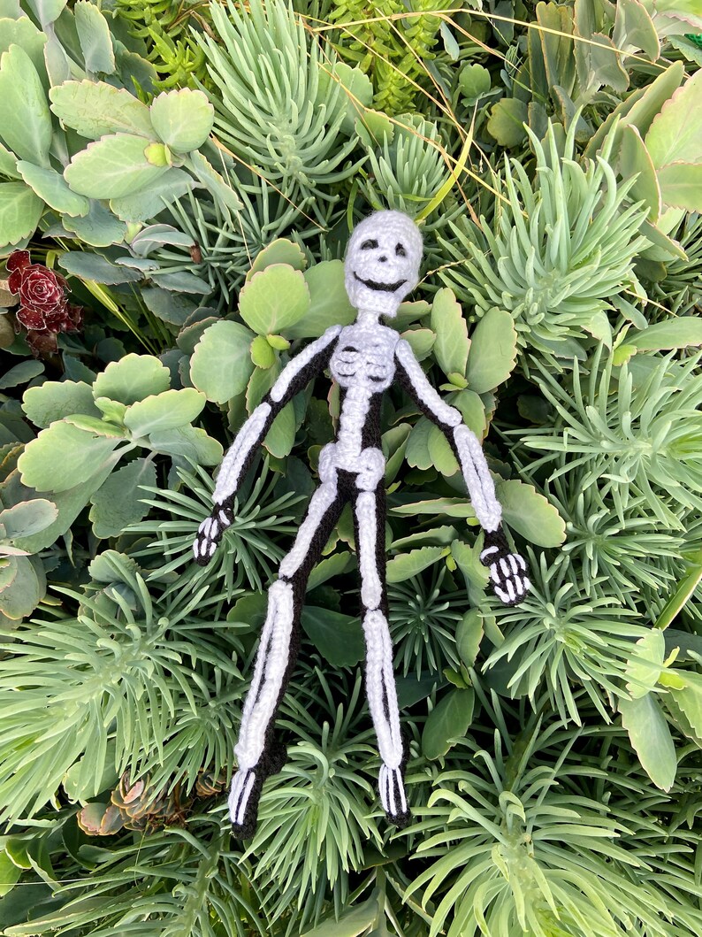 May include: A crocheted skeleton toy with a smiling face, lying on a bed of green succulents.