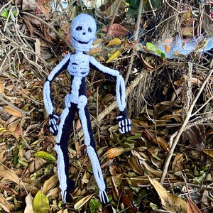 May include: A crocheted skeleton doll with a white body and black limbs, standing in a bed of brown leaves and twigs.