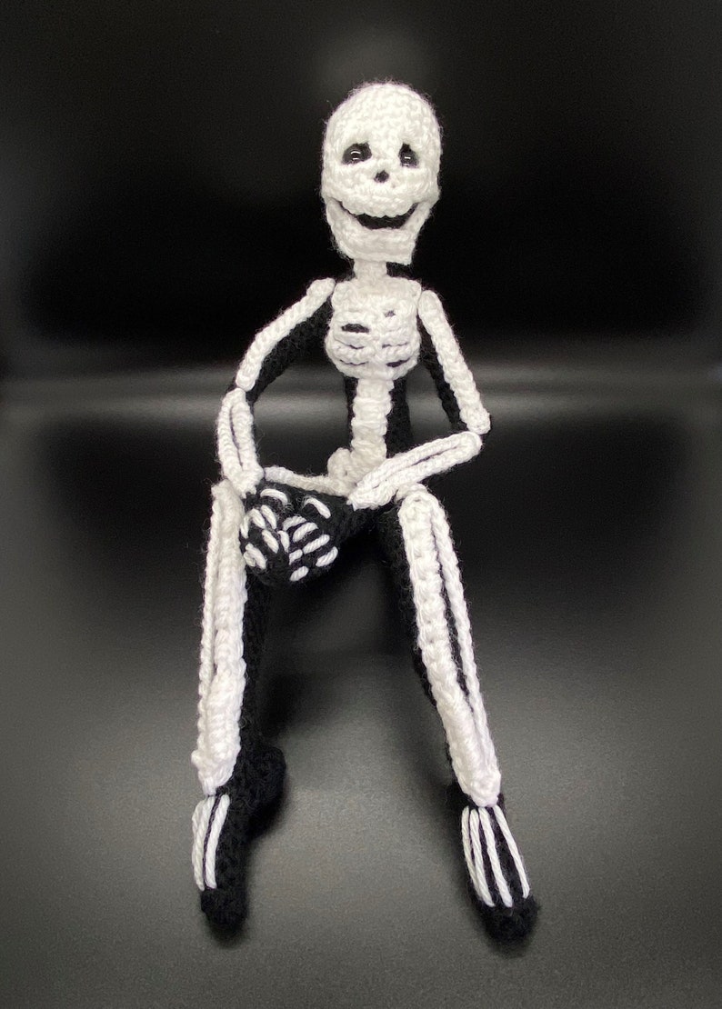 May include: A crocheted skeleton doll with a white skull and black body. The skeleton is sitting with its legs crossed and its hands clasped in front of it.