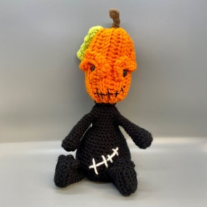 May include: A crocheted pumpkin-headed doll with a black body and white stitching. The doll is sitting on a gray surface.