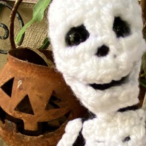 May include: A white crocheted skeleton doll with black eyes and a friendly smile. The doll is wearing a black and white striped shirt. The doll is sitting in front of a rusty metal pumpkin.