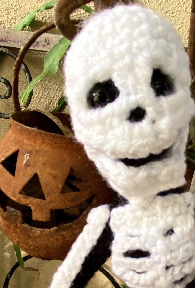 May include: A white crocheted skeleton doll with black eyes and a friendly smile. The doll is wearing a black and white striped shirt. The doll is sitting in front of a rusty metal pumpkin.