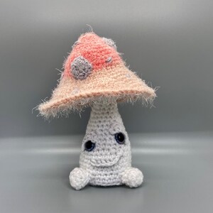 May include: A crocheted mushroom toy with a pink and white cap and a white body. The mushroom has a smiling face with black eyes and a white stem.
