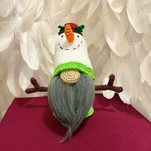 May include: A handmade snowman gnome with a white crocheted body, green and beige accents, and a gray beard. It has a carrot nose, twig arms, and a festive hat with holly and berries. The whimsical design is perfect for holiday decor.