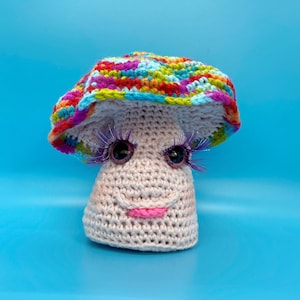 May include: A crocheted mushroom with a colorful hat and large eyelashes. The mushroom is pink and has a smiling face.