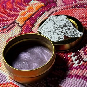 May include: A small, open gold-colored tin containing shimmering, lavender-colored putty. The lid, also gold, features a black and white illustration of grapes and leaves, with the text "Grape AB Putty". The background is a colorful, patterned surface.