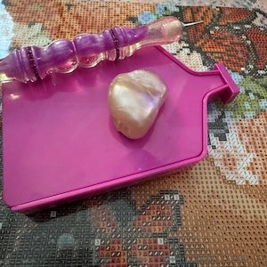 May include: A pink diamond painting tray with a pearlescent bead and a purple diamond painting pen. The pen has a clear, glittery tip and a purple handle. The tray is rectangular with a pouring spout.
