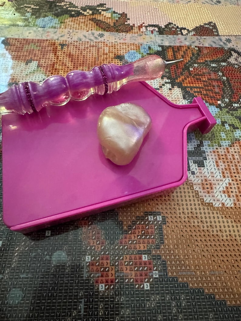 May include: A pink diamond painting tray with a pearlescent bead and a purple diamond painting pen. The pen has a clear, glittery tip and a purple handle. The tray is rectangular with a pouring spout.