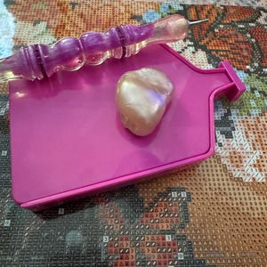 May include: A pink diamond painting tray with a pearlescent stone and a purple and clear diamond painting pen. The tray is rectangular with a small handle. The pen has a purple, glittery handle.