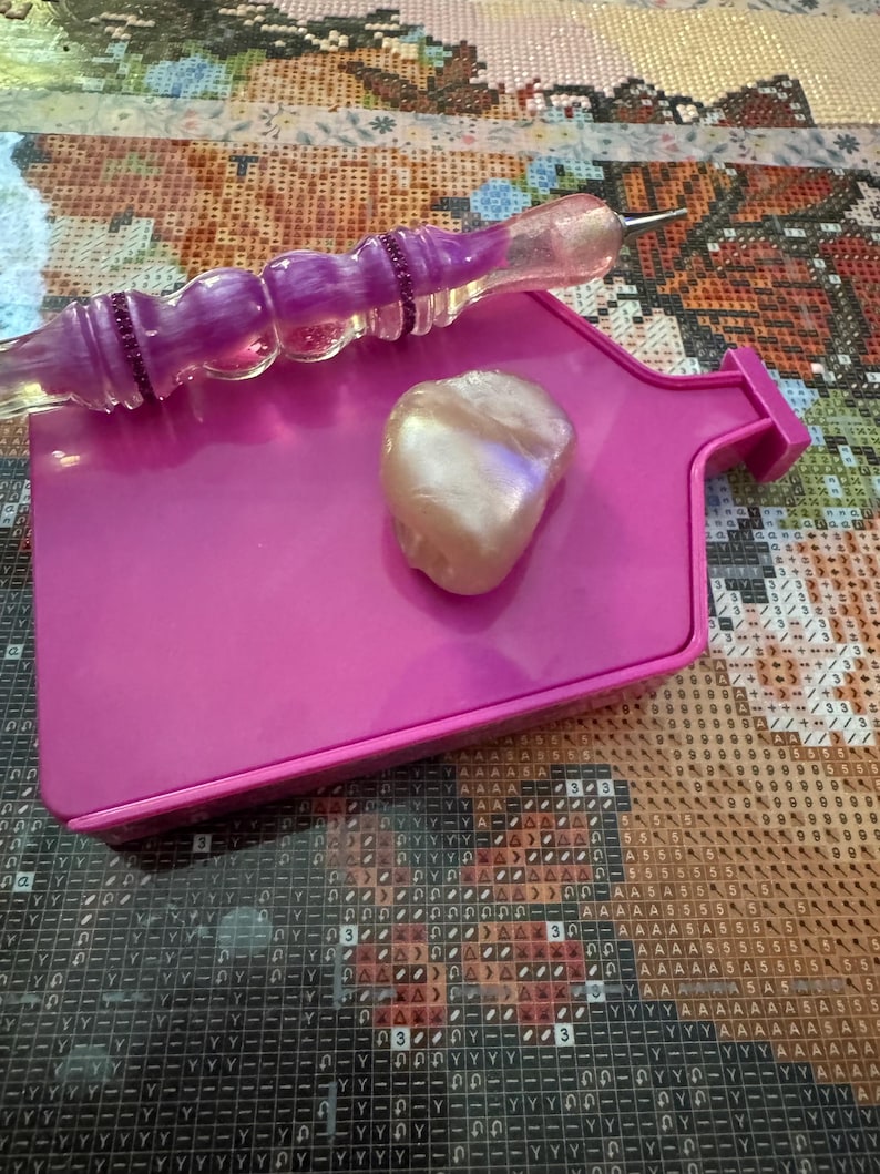 May include: A pink diamond painting tray with a pearlescent stone and a purple and clear diamond painting pen. The tray is rectangular with a small handle. The pen has a purple, glittery handle.