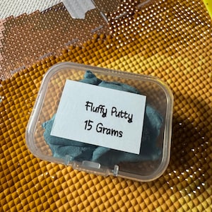 May include: A clear plastic container holding teal-colored fluffy putty. A white label inside the container reads "Fluffy Putty 15 Grams". The putty is a soft, pliable material, perfect for crafts and sensory play.