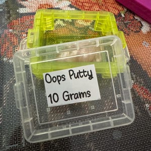 May include: Clear plastic container with a neon green lid, labeled "Oops Putty 10 Grams." The container is rectangular with a hinged lid and is partially filled with a light-colored putty. The container is sitting on a patterned surface.