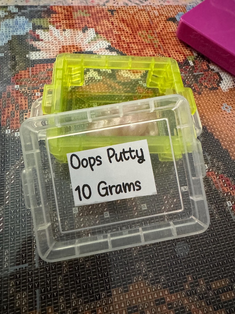 May include: Clear plastic container with a neon green lid, labeled "Oops Putty 10 Grams." The container is rectangular with a hinged lid and is partially filled with a light-colored putty. The container is sitting on a patterned surface.