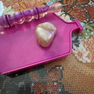 May include: A pink diamond painting tray with a purple and clear stylus and a pearlescent wax square. The stylus has a decorative handle with purple accents. The tray is rectangular with a raised edge.