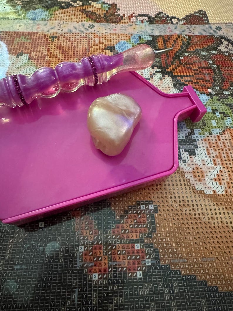 May include: A pink diamond painting tray with a purple and clear stylus and a pearlescent wax square. The stylus has a decorative handle with purple accents. The tray is rectangular with a raised edge.