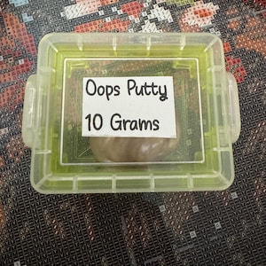 May include: A clear, rectangular container with a green frame holds a small amount of brown putty. A white label inside the container reads "Oops Putty 10 Grams". The container is on a patterned surface.