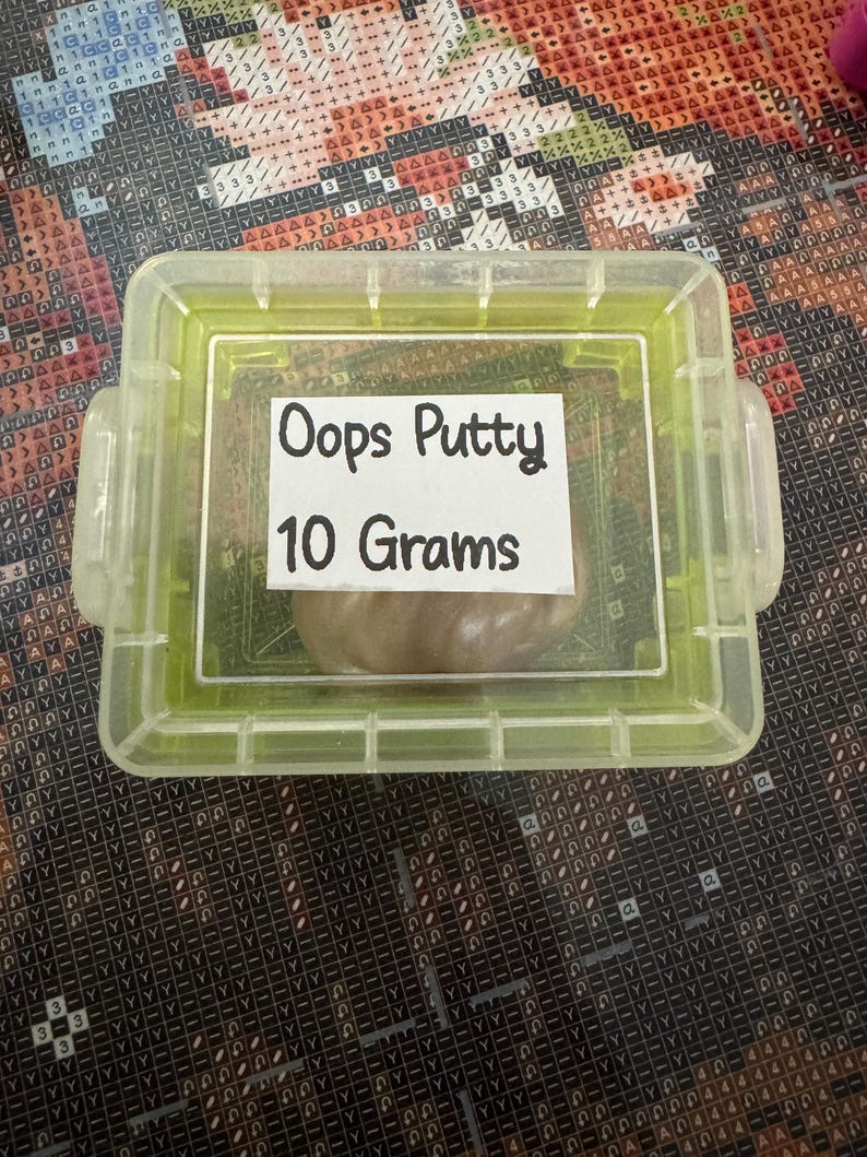 May include: A clear, rectangular container with a green frame holds a small amount of brown putty. A white label inside the container reads "Oops Putty 10 Grams". The container is on a patterned surface.