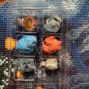 May include: A clear plastic organizer containing six small, colorful wax-like substances. The colors include shades of blue, orange, gray, and a tan-yellow. The items are likely for a craft project, such as diamond painting.