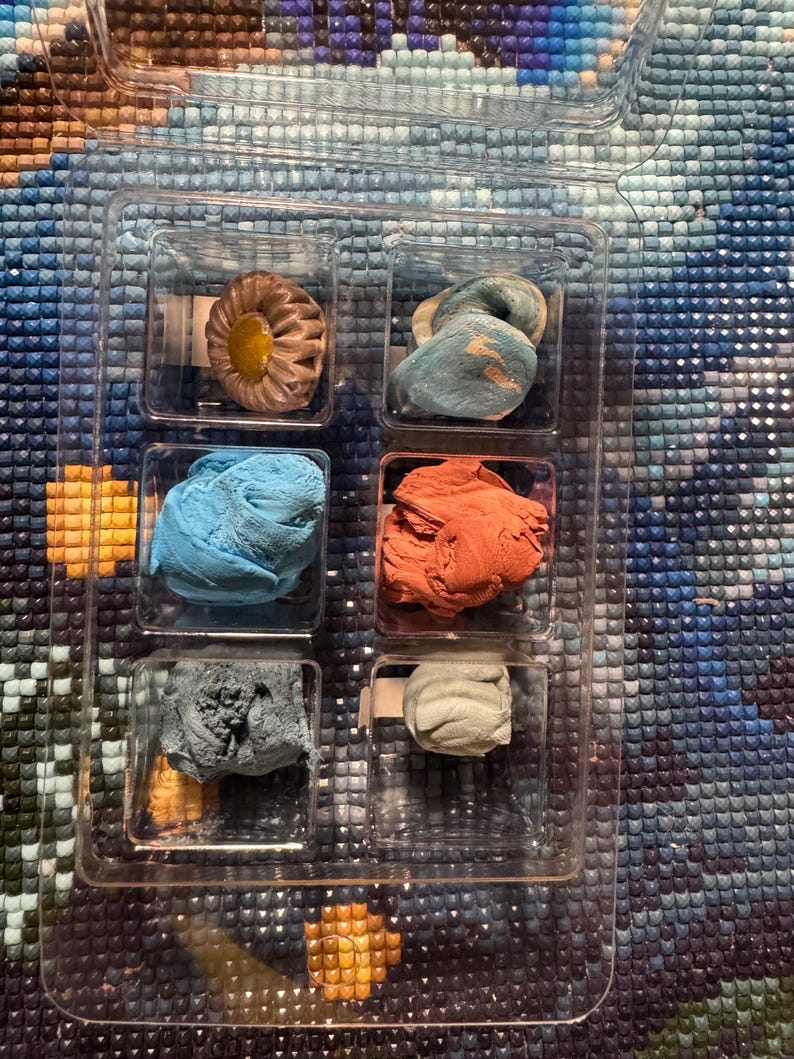 May include: A clear plastic organizer containing six small, colorful wax-like substances. The colors include shades of blue, orange, gray, and a tan-yellow. The items are likely for a craft project, such as diamond painting.