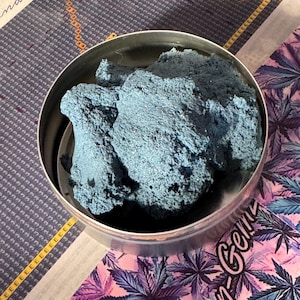May include: A round, silver container filled with a large mass of blue material. The material has a textured, granular appearance. The container sits on a patterned surface with text and a floral design in pink and purple.