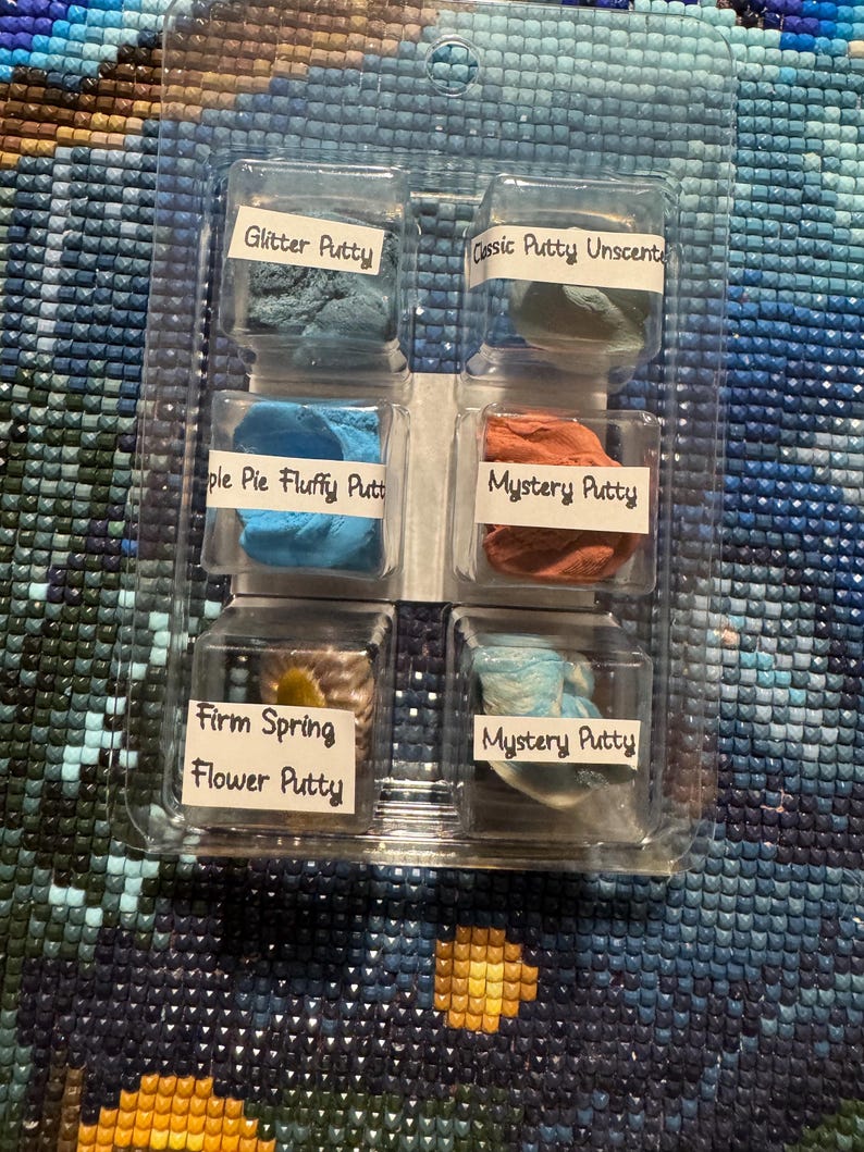 May include: A clear plastic container holding six small containers of colorful putty. The putty colors include blue, green, orange, and a mix of blue and white. Each container is labeled with a different name, such as "Glitter Putty" and "Mystery Putty".