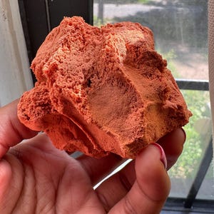 May include: A raw, porous, and irregularly shaped chunk of terracotta-colored clay. The surface exhibits a rough, granular texture with visible indentations and a matte finish, showcasing a deep, earthy orange-red hue throughout the entire piece.