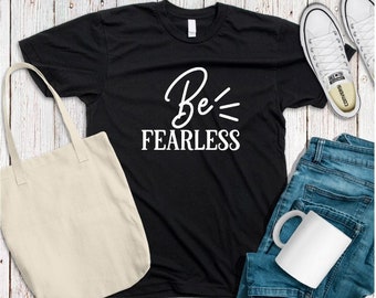 以下が含まれることがあります： 白い文字で「Be Fearless」と書かれた黒いTシャツ。Tシャツは、白いトートバッグ、白いスニーカー、白いマグカップと一緒に、木製の表面に平らに置かれています。