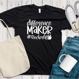 Op de afbeelding: Zwart T-shirt met witte tekst die "difference maker #teacherlife" luidt en een rode appelafbeelding. Het shirt ligt op een houten oppervlak met een witte tote bag, een blauwe spijkerbroek en een witte mok.