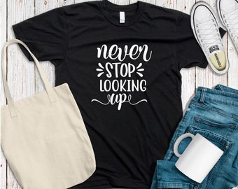 May include: Black t-shirt with white text that reads "never stop looking up". The shirt is laid out on a wooden surface with a tote bag, a mug, and a pair of jeans.