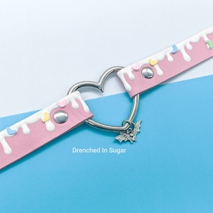 May include: Pink faux leather choker with white icing drip and colorful sprinkles. A silver heart-shaped clasp with a bat charm hangs from the choker. The text "Drenched In Sugar" is printed below the choker.