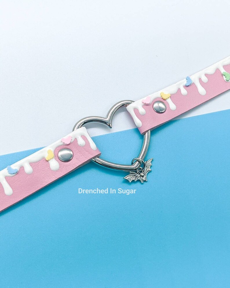 May include: Pink faux leather choker with white icing drip and colorful sprinkles. A silver heart-shaped clasp with a bat charm hangs from the choker. The text "Drenched In Sugar" is printed below the choker.