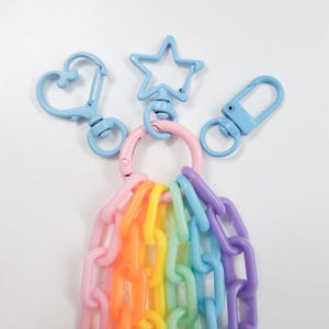 May include: A rainbow chain keychain with a light blue heart, star, and rectangle shaped charms. The charms are attached to a pink ring.