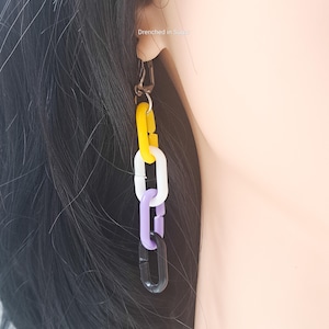 May include: A dangling earring featuring a series of colorful chain links. The links are yellow, white, purple, and black. The earring is attached to a silver clasp. The text "Drenched in Sugar" is visible in the background.