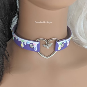 May include: Purple choker necklace with a heart-shaped charm and a bat pendant. The choker is decorated with white and pastel-colored drips and sprinkles. The text "Drenched in Sugar" is visible in the image.